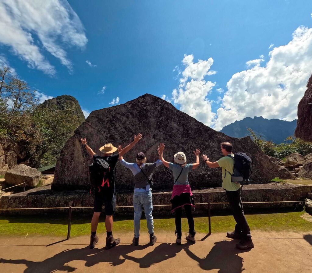 A group of four travelers stands with arms raised in front of the Sacred Rock inside the Machu Picchu citadel – Peru Hikers