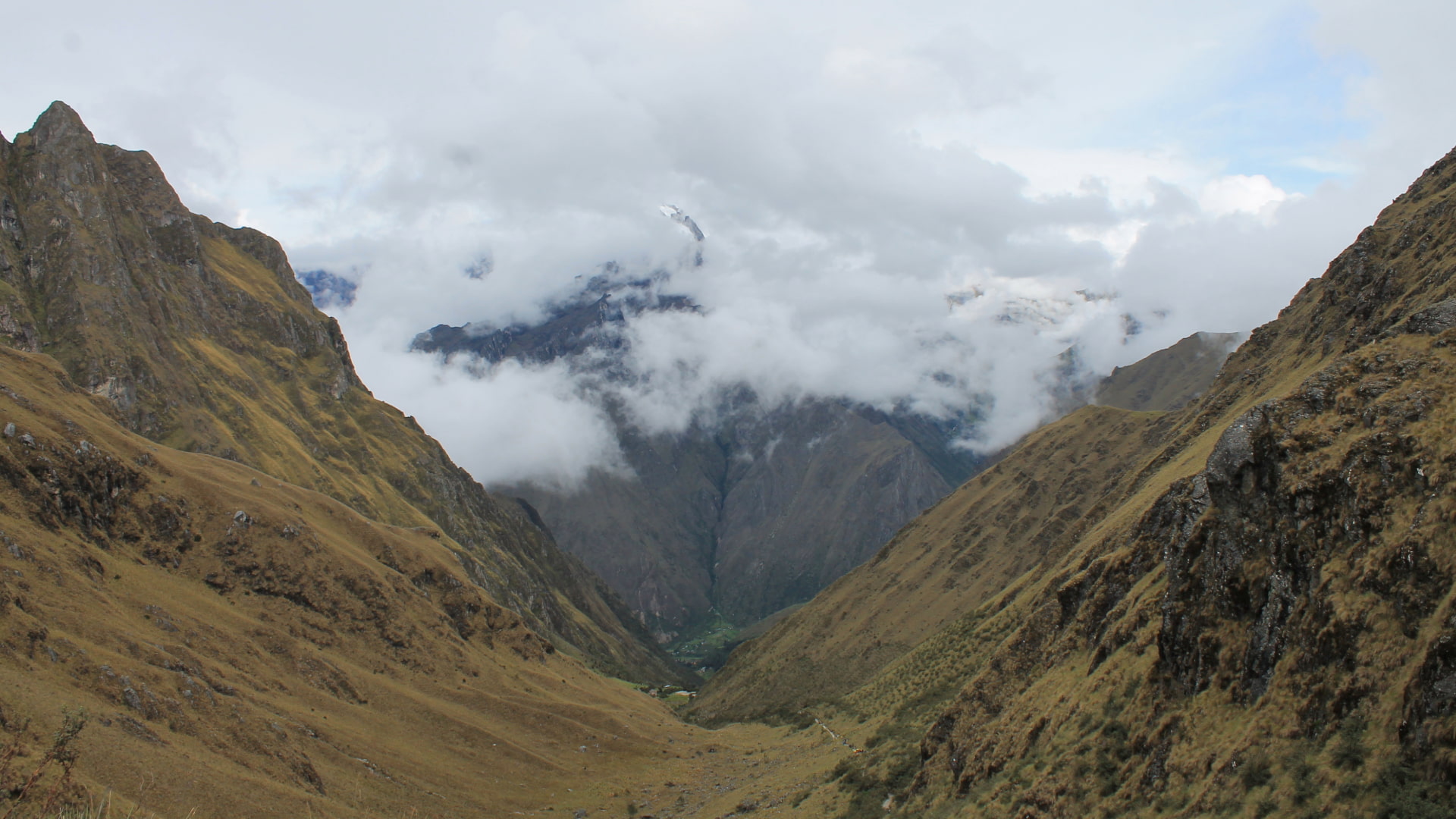 View from the top of Dead Woman's Pass on the Inca Trail, looking down into a deep Andean valley with cloud-covered peaks in the distance - Peru Hikers.